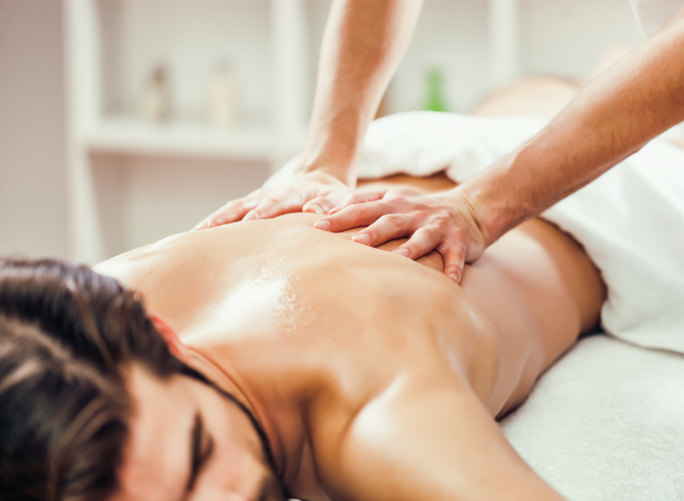 Ein Mann liegt oberkörperfrei auf dem Bauch und wird auf dem Rücken von zwei Händen massiert. Seinen Kopf hat er leicht zur Seite geneigt abgelegt.