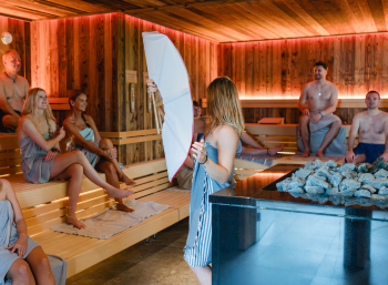 Mehrere Personen sitzen verteilt in einer Saunakabine, während eine junge Frau vor dem Ofen mit einem Saunafächer wedelt.